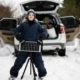 Boy with solar panel battery on tripod against car in winter woods.