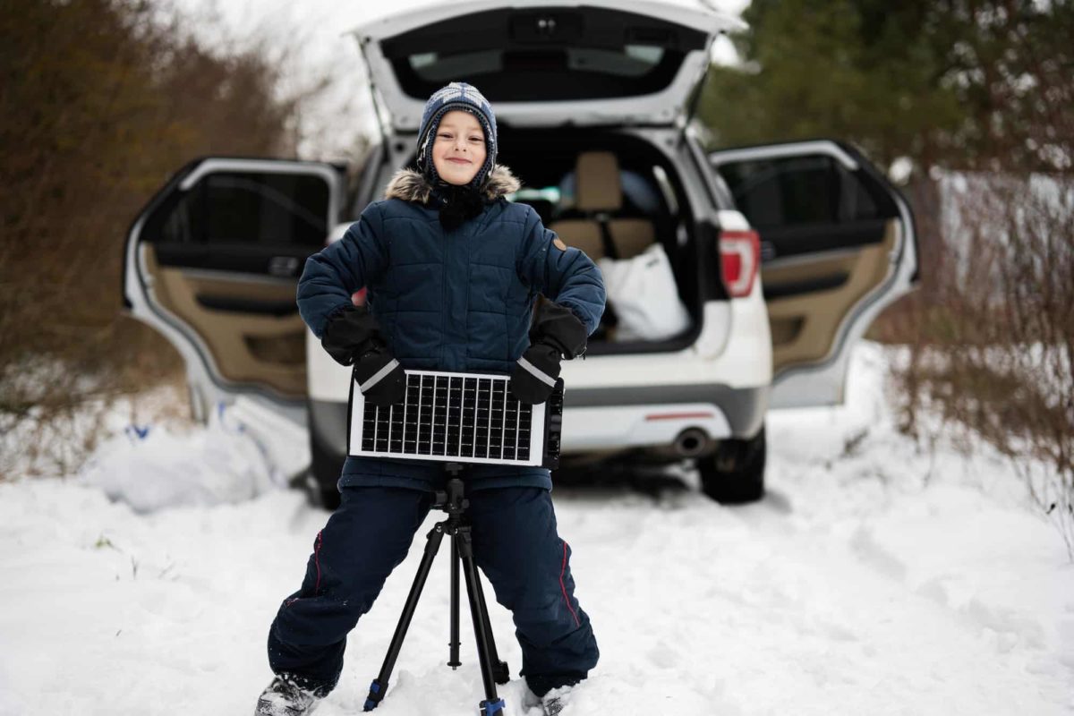 Boy with solar panel battery on tripod against car in winter woods.